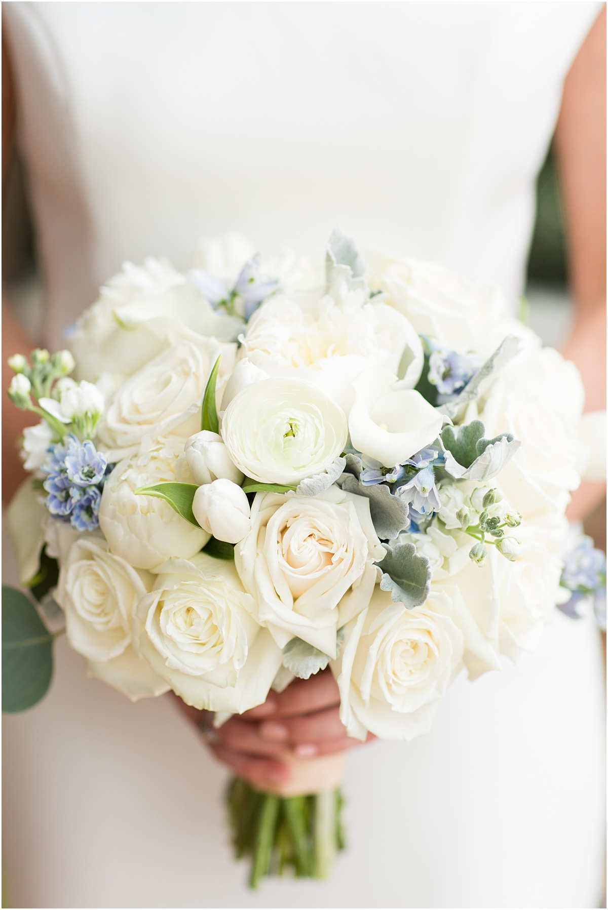 Bride holding a white bridal bouquet with roses, ranunculus and blue flowers.